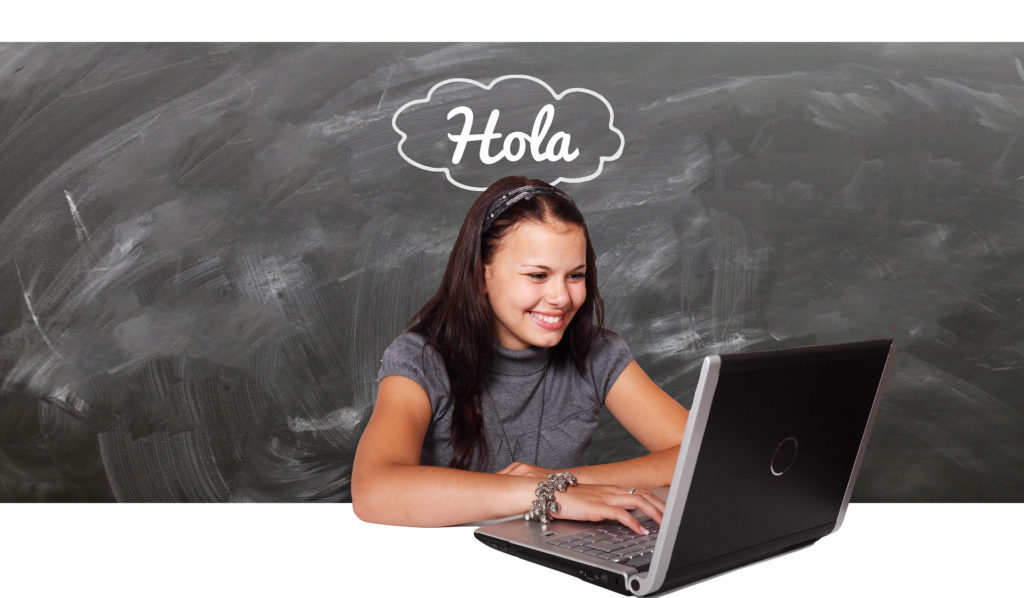woman at a laptop in front of a chalkboard that says, "hola"